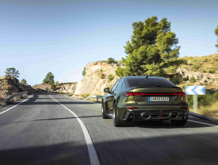 Achteraanzicht van de rijdende Audi RS5 in het groen op een zonnige, uitgestrekte weg.