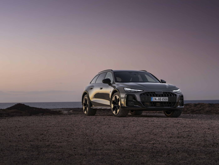 Donkergrijze Audi A6 Allroad stationwagon vooraanzicht bij zonsondergang aan de kustlijn.