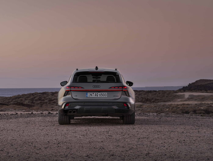 Achteraanzicht van de donkergrijze Audi A6 Allroad stationwagon tegen een zonsondergang aan zee.