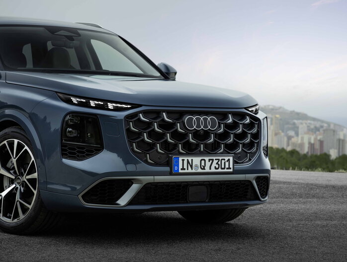 Close-up van Audi Q7's gedetailleerde grille en koplampen, met stadslandschap op afstand.