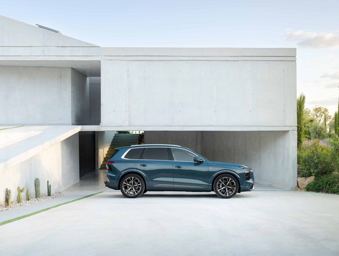Petrolblauwe Audi Q7 SUV, zijkant, geparkeerd naast modern architectonisch gebouw in de zon.
