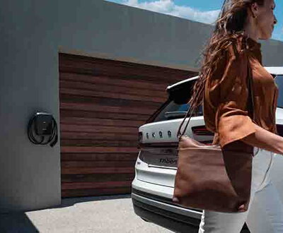 Witte Škoda Kodiaq op oprit met vrouw die voorbij stapt. In de achtergrond zie je een wallbox laadstation aan de muur hangen