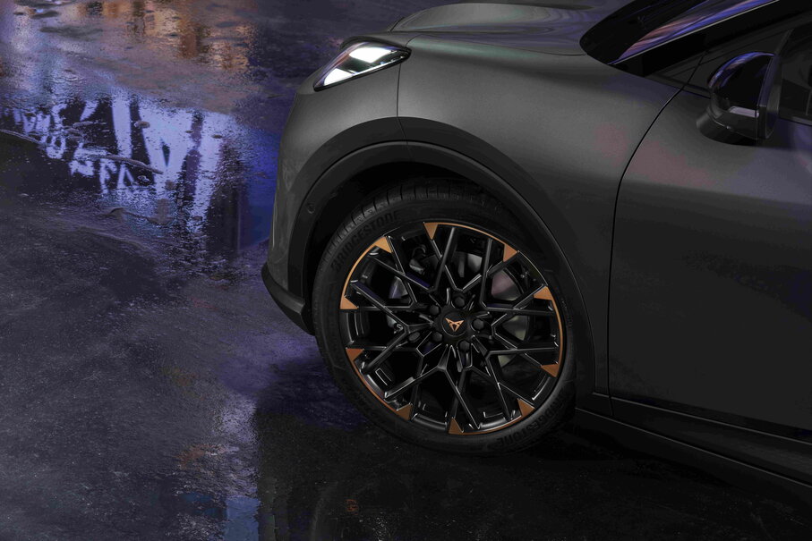De donkergrijze Cupra Raval toont zijn sportieve velgen met bronzen accenten op een natte ondergrond.