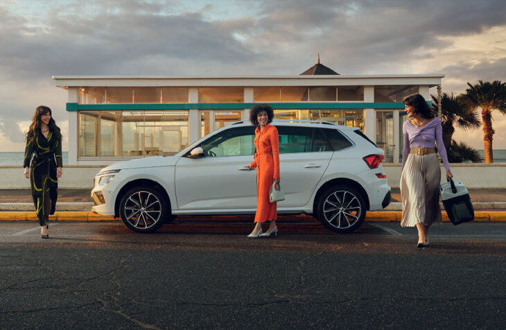 Witte Škoda Kamiq geparkeerd aan zee tijdens zonsondergang, met drie vriendinnen die voorbij wandelen in een moderne lifestyle-setting