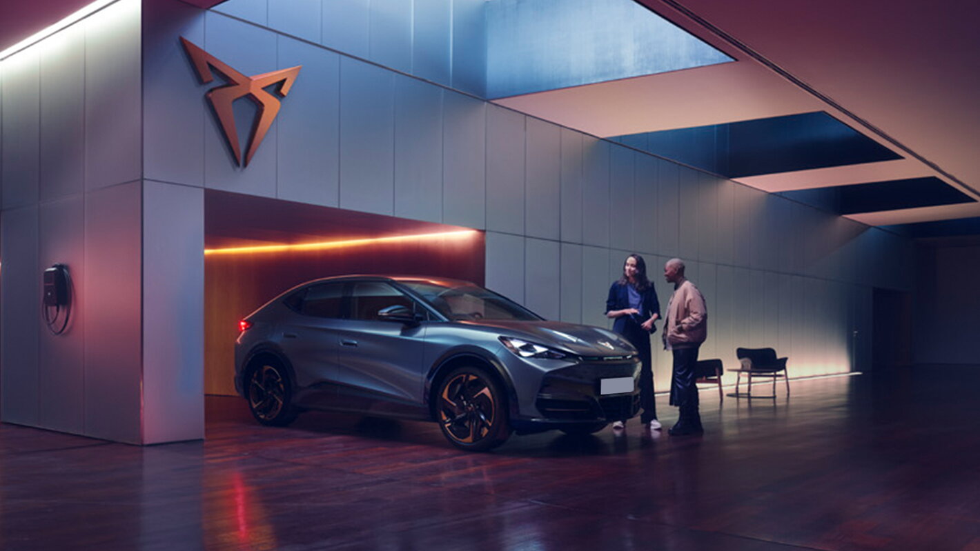 De elektrische Cupra Tavascan SUV in een moderne showroom met sfeerverlichting en een laadstation aan de wand.