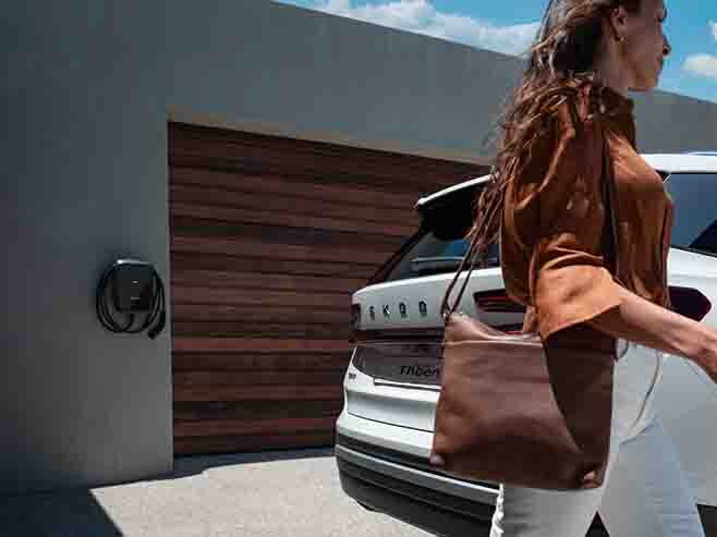 Witte Škoda Kodiaq op oprit met vrouw die voorbij stapt. In de achtergrond zie je een wallbox laadstation aan de muur hangen