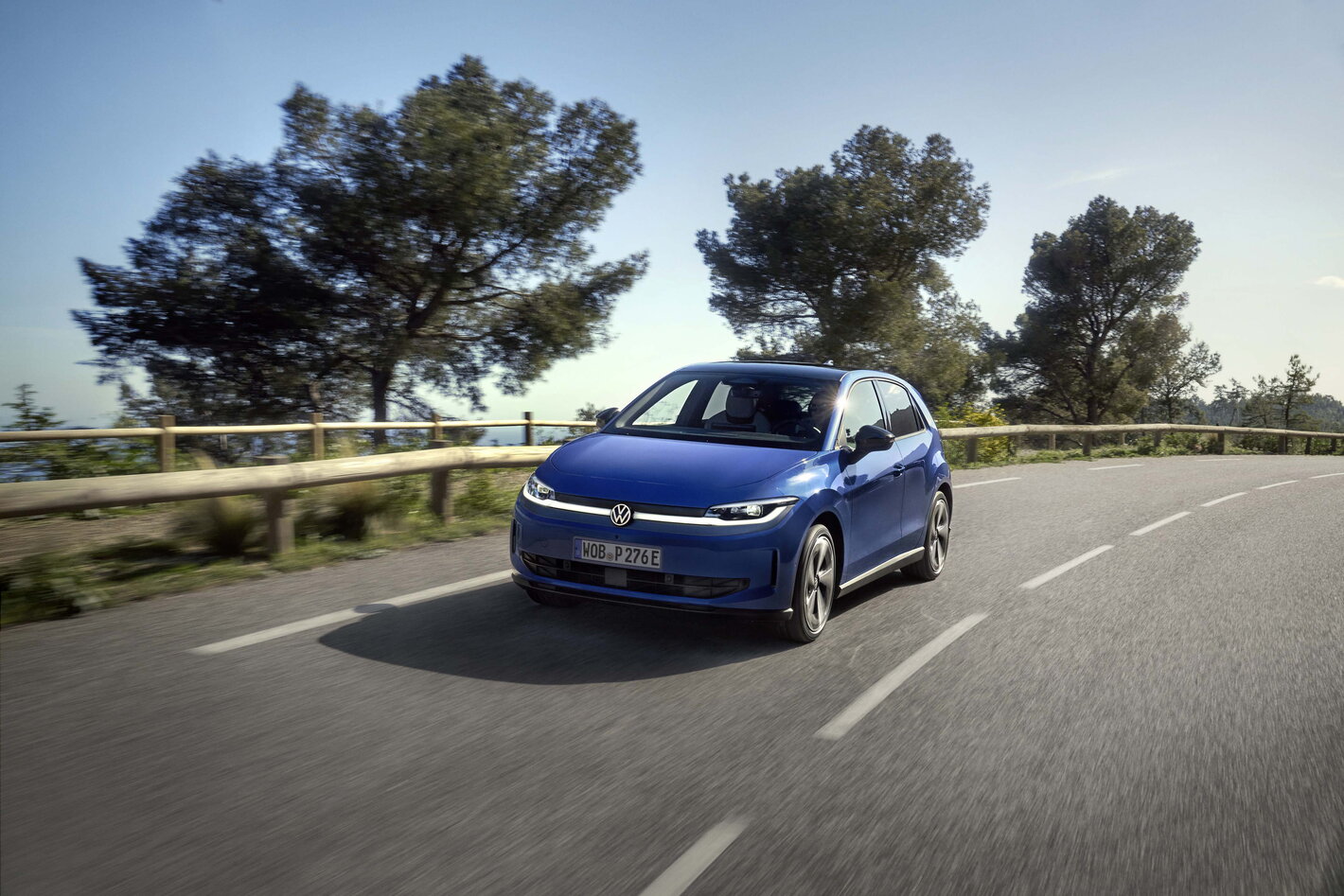 Blauwe Volkswagen Id.Polo rijdt dynamisch over een kronkelende weg met bomen en een blauwe lucht.