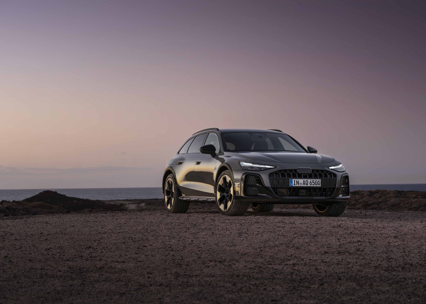 Donkergrijze Audi A6 Allroad stationwagon vooraanzicht bij zonsondergang aan de kustlijn.