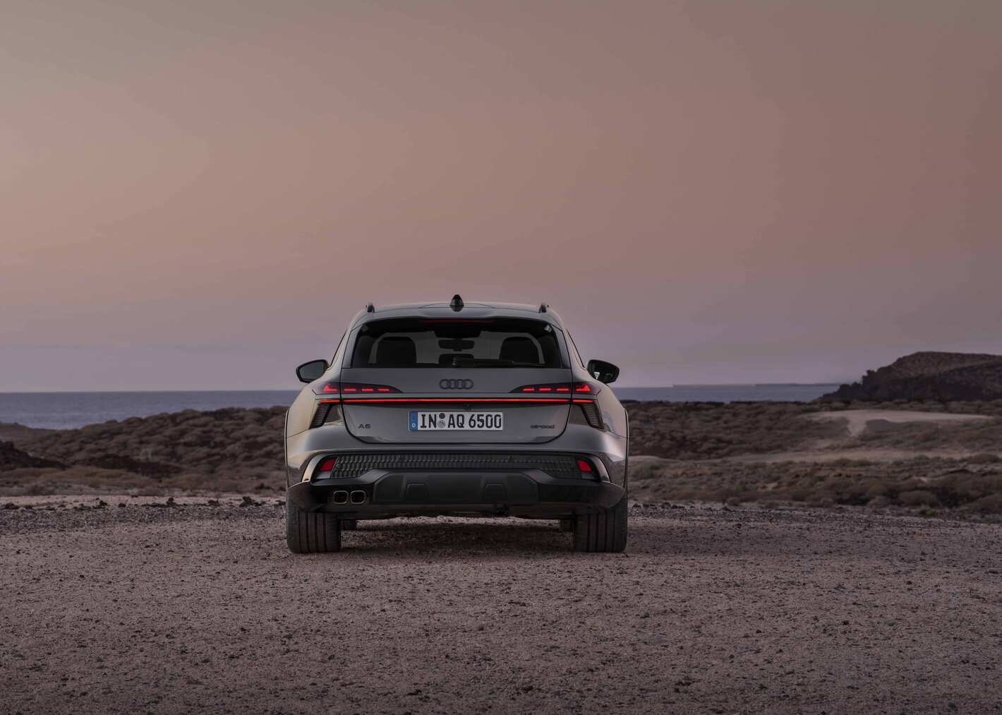 Achteraanzicht van de donkergrijze Audi A6 Allroad stationwagon tegen een zonsondergang aan zee.
