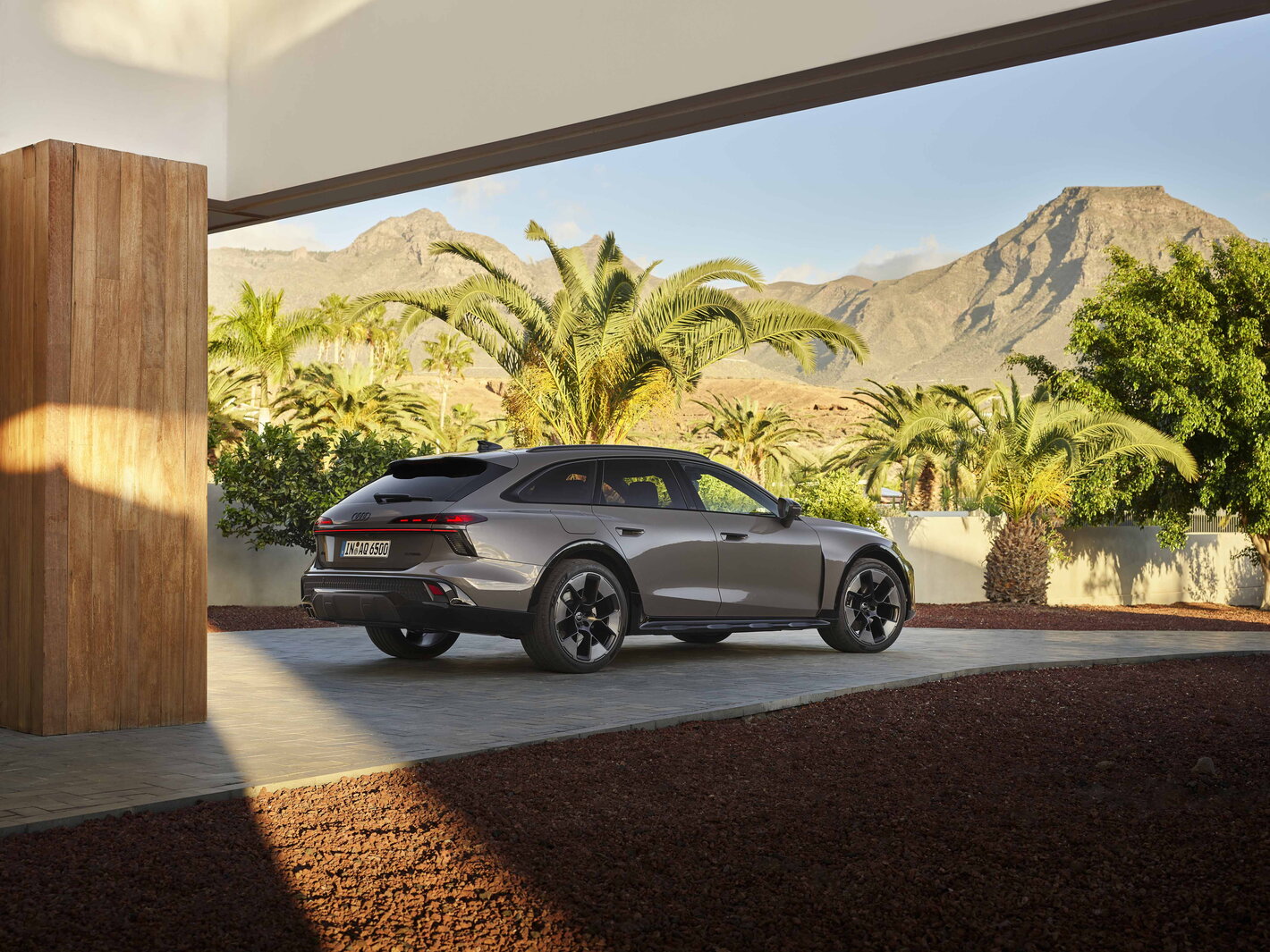 Grijze Audi A6 Allroad geparkeerd bij luxe villa met palmbomen en bergen.